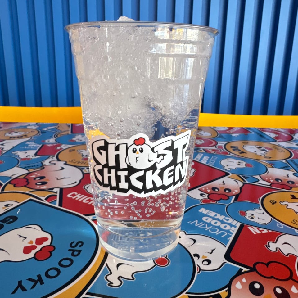 A clear Ghost Chicken branded cup filled with sparkling soda and ice on a Ghost Chicken patterned table.