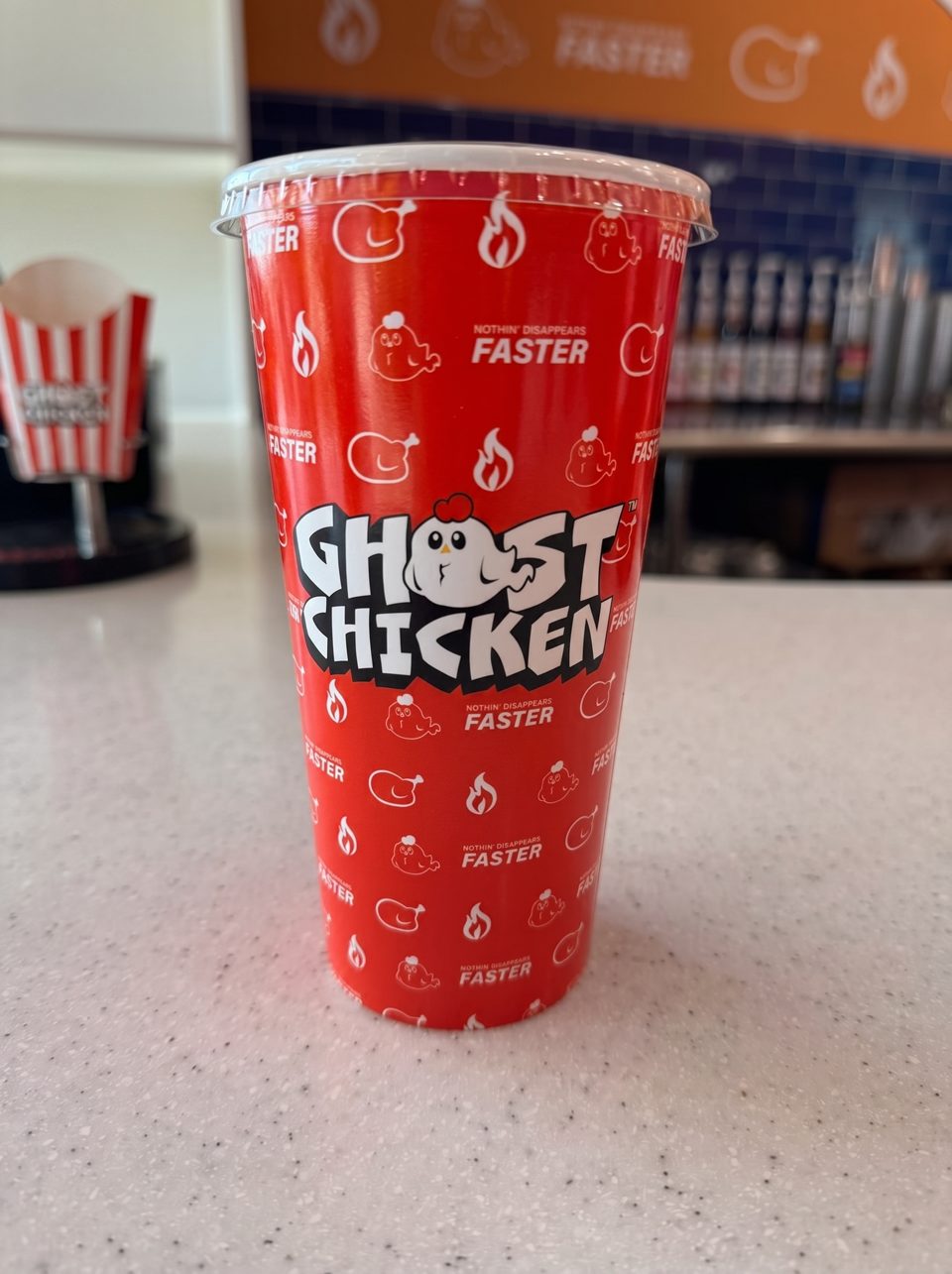 A red Ghost Chicken branded cup with a white lid sits on a speckled white counter.